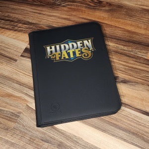 May include: Black zippered binder with a gold and blue "HIDDEN FATES" logo. The binder has a small white logo that says "Ultra Pro".