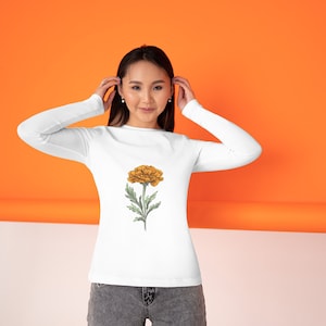 May include: White long-sleeve shirt featuring a detailed illustration of a yellow flower with green leaves and stem. The floral design is centered on the front of the shirt. The background is orange and white.