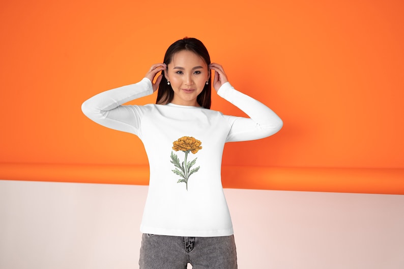 May include: White long-sleeve shirt featuring a detailed illustration of a yellow flower with green leaves and stem. The floral design is centered on the front of the shirt. The background is orange and white.