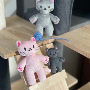 May include: Three plush cat toys in gray, pink, and dark gray, each with embroidered facial features and pink accents on the ears and paws. A blue ball of yarn sits near the gray cat. The toys are on a wooden cat tree.