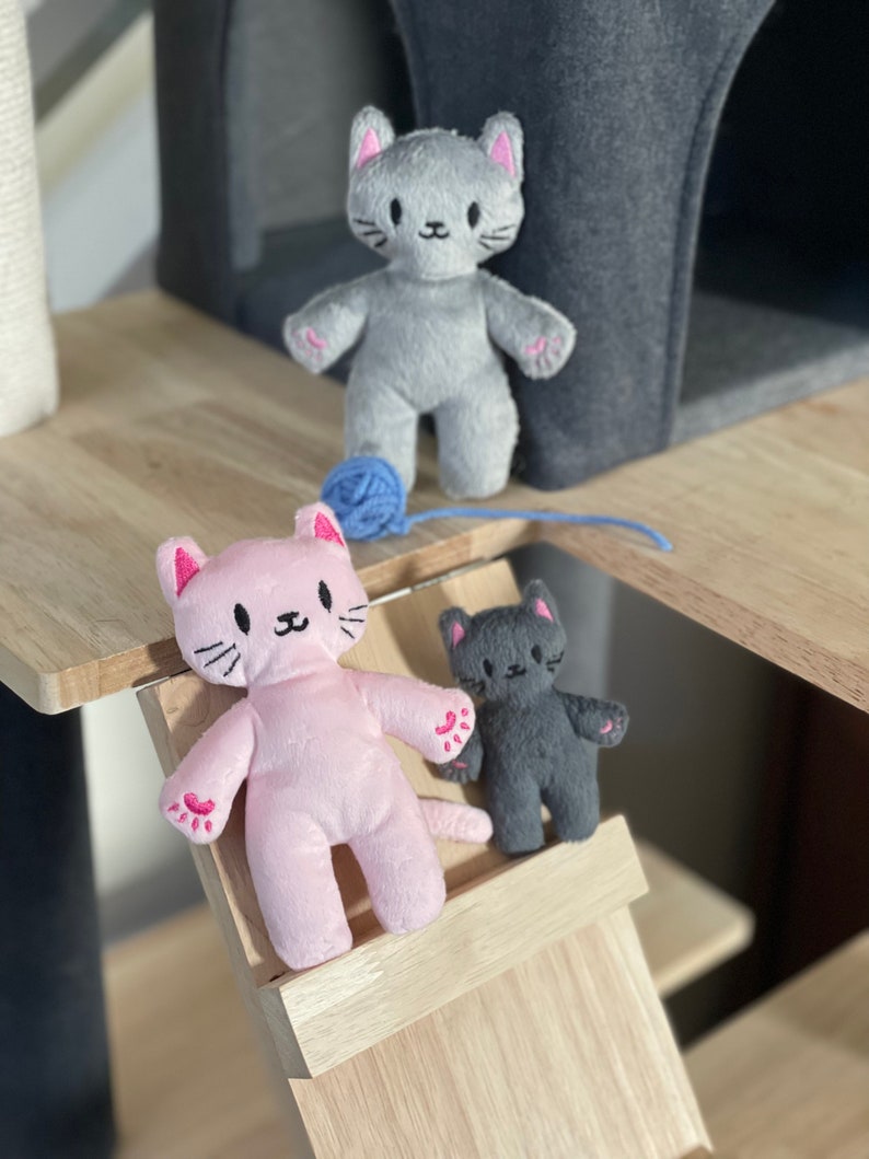 May include: Three plush cat toys in gray, pink, and dark gray, each with embroidered facial features and pink accents on the ears and paws. A blue ball of yarn sits near the gray cat. The toys are on a wooden cat tree.