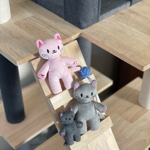 May include: A cat tree with plush cat toys. The toys are in pink, gray, and dark gray. The cat toys are on a wooden ramp. A blue yarn ball is next to the pink cat toy. The cat tree has a gray felted cat house.