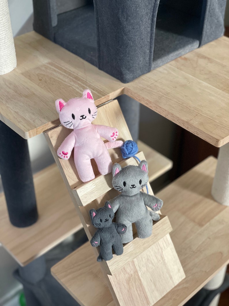 May include: A cat tree with plush cat toys. The toys are in pink, gray, and dark gray. The cat toys are on a wooden ramp. A blue yarn ball is next to the pink cat toy. The cat tree has a gray felted cat house.