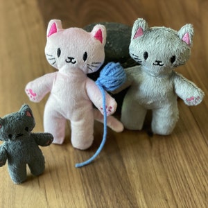 May include: Three plush cat toys in pink, gray, and dark gray. The pink cat holds a blue yarn ball. Each cat has embroidered details, including pink inner ears and paw pads. The toys are on a wooden surface.