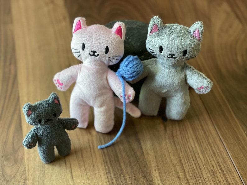 May include: Three plush cat toys in pink, gray, and dark gray. The pink cat holds a blue yarn ball. Each cat has embroidered details, including pink inner ears and paw pads. The toys are on a wooden surface.