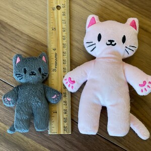 May include: Two plush cat toys, one gray and one pink, with embroidered facial features and paw details. The pink cat is approximately 10 inches tall. A wooden ruler is placed next to the toys for scale.
