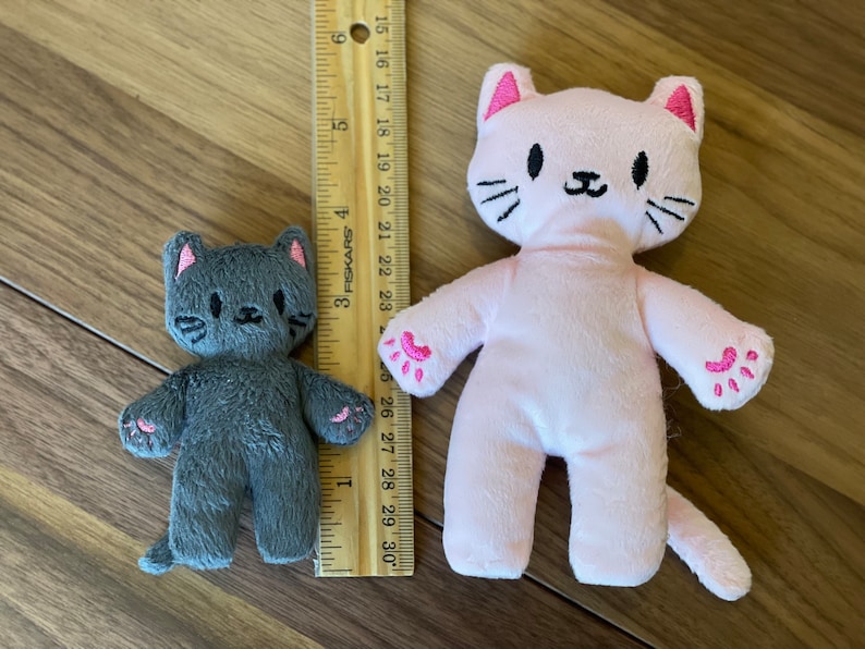 May include: Two plush cat toys, one gray and one pink, with embroidered facial features and paw details. The pink cat is approximately 10 inches tall. A wooden ruler is placed next to the toys for scale.