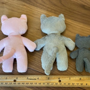 May include: Three plush cat toys in varying sizes and colors: pink, gray, and dark gray. Each cat has a tail and is positioned with its back facing the viewer. A ruler is placed below the toys for size comparison.