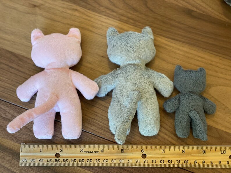 May include: Three plush cat toys in varying sizes and colors: pink, gray, and dark gray. Each cat has a tail and is positioned with its back facing the viewer. A ruler is placed below the toys for size comparison.