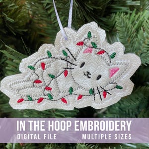 May include: White cat Christmas ornament embroidered with a festive design. The cat is wrapped in a string of red and green Christmas lights. The ornament is hanging on a Christmas tree. Text on the image reads "IN THE HOOP EMBROIDERY DIGITAL FILE MULTIPLE SIZES."