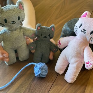 May include: Three plush cat toys in gray and pink, with embroidered facial features and pink paw pads. A blue yarn ball sits near the toys. The toys are on a wooden surface.