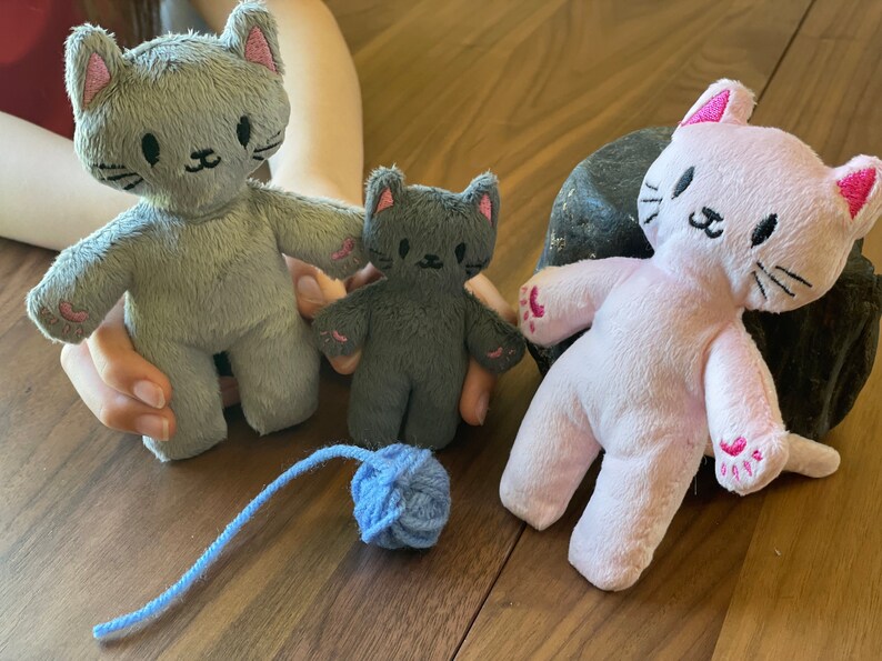 May include: Three plush cat toys in gray and pink, with embroidered facial features and pink paw pads. A blue yarn ball sits near the toys. The toys are on a wooden surface.