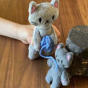 May include: Two gray plush cat dolls with pink accents. One doll holds a ball of blue yarn, while the other sits nearby. The dolls have embroidered facial features and are made of a soft, textured material. The dolls are on a wooden surface.