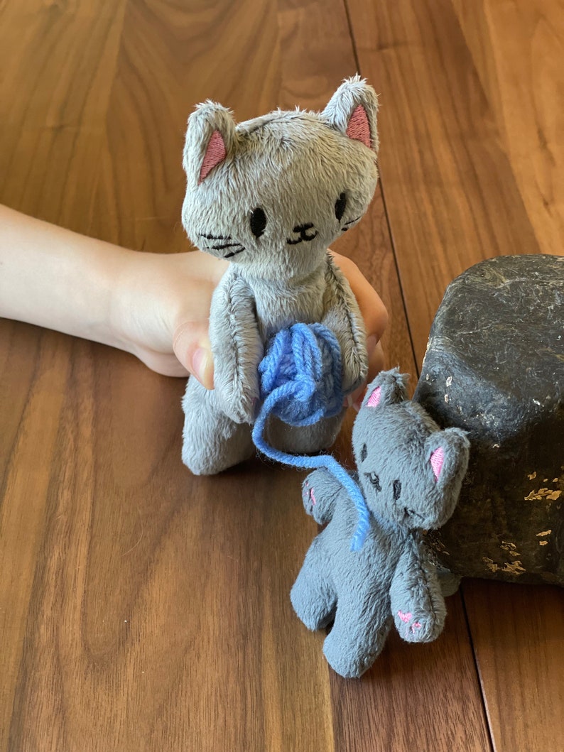 May include: Two gray plush cat dolls with pink accents. One doll holds a ball of blue yarn, while the other sits nearby. The dolls have embroidered facial features and are made of a soft, textured material. The dolls are on a wooden surface.