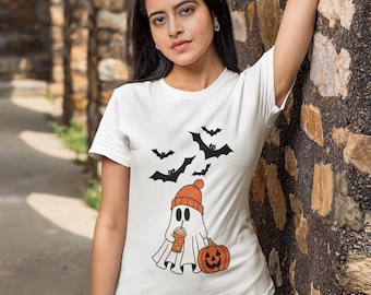 May include: White t-shirt featuring a Halloween-themed graphic. The design includes a ghost wearing an orange beanie and holding a drink, a carved pumpkin, and several black bats. The shirt is paired with khaki pants.