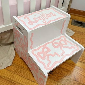 May include: A white two-step stool with pink bow accents and the name "Leighton" painted on top. The step stool is designed for children and is placed on a hardwood floor.