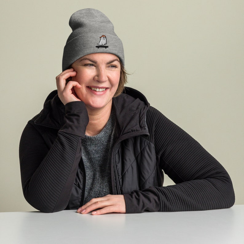 May include: A woman wearing a gray beanie with a white ghost skateboarding graphic. She is smiling and wearing a black jacket.
