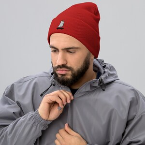 May include: A red beanie with a white embroidered ghost wearing a red hat and riding a skateboard.