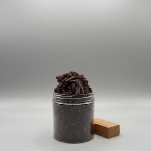 May include: A clear plastic jar filled with dark red, shredded material, possibly wood chips or a similar substance. A small, rectangular wooden block rests beside the jar. The background is a plain, light gray.
