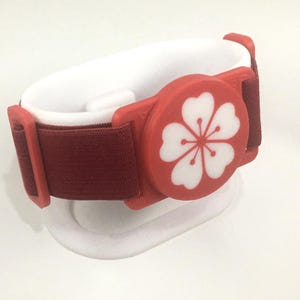 Puede incluir: Una banda elástica de color granate con un componente circular de plástico rojo que presenta un diseño de flor blanca de cinco pétalos. La banda está asegurada por deslizadores de plástico rojo a cada lado, creando un conjunto de correa funcional.