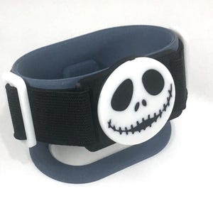 May include: A black and blue wristband featuring a white Jack Skellington face. The wristband has a hook and loop closure and a blue loop. The Jack Skellington face is a circular design with black eyes and a stitched mouth.