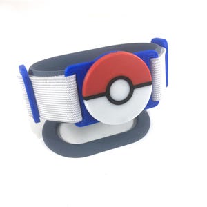 May include: A gray, white, red, and blue wristband featuring a circular design reminiscent of a Poké Ball. The wristband has a white strap and a gray base, with blue accents and a red and white circular emblem.