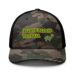 Puede incluir: Gorra de camionero de camuflaje con parte trasera de malla negra y visera curva. La parte delantera presenta el texto bordado "BLOOPBLOOP YBYSAIA" en dorado, junto con un gráfico de tortuga verde. El sombrero es una mezcla de verde, marrón y negro.