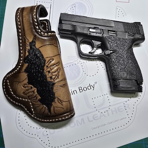 May include: A brown leather holster with a black design, made for a small black handgun. The holster is laid out on a white sheet of paper with a pattern of small holes. The text "Copyright 2024 - JDub Custom Leather" is printed on the paper.
