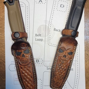 Peut inclure: Deux couteaux personnalisés avec étuis en cuir. L'un a un manche beige, l'autre noir. Les étuis en cuir marron présentent des motifs de crânes sculptés. Les couteaux sont présentés sur un papier avec un motif et le texte 'Belt Loop'.