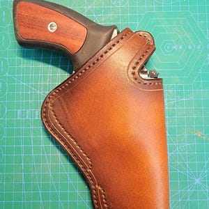 以下が含まれることがあります： 銃器を収納するために設計された、ステッチデザインのついた茶色の革製ホルスター。ホルスターは緑色のカッティングマットの上に置かれています。