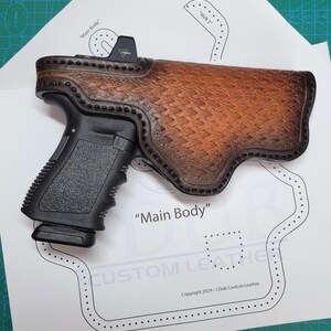 Puede incluir: Una pistola negra insertada en una funda de cuero marrón hecha a medida con un patrón de tejido de cesta y bordes cosidos oscuros. La funda descansa sobre una plantilla de papel blanco con las etiquetas Main Body, Welt 2 y Copyright 2024.