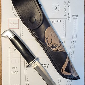 May include: A black leather knife sheath with an alien skull design. The sheath is lying on a white piece of paper with a pattern for making a leather sheath. A silver and black handled knife is lying on the paper.