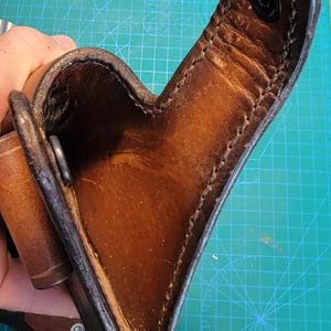 以下が含まれることがあります： ステッチのディテールと金属製の留め具が付いた茶色の革製ホルスター。ホルスターは開いており、空です。