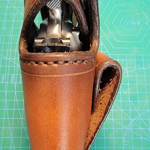 以下が含まれることがあります： 銃器用の茶色の革製ホルスター。ホルスターは銃の上部を覆うフラップと、側面にベルトループが付いています。