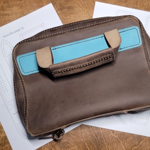 May include: A brown leather zippered pouch with a blue and tan leather handle. The pouch is sitting on a white sheet of paper with a pattern for a leather strap.