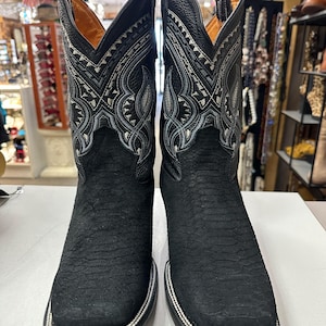 May include: A pair of black cowboy boots with intricate white and gray stitching on the upper shaft. The boots have a textured leather appearance and a square toe design. The soles are outlined with a silver trim.