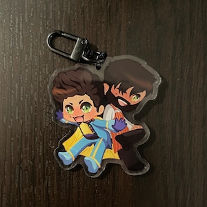 May include: A clear acrylic keychain featuring a cartoon illustration of two people. One person is wearing a blue and yellow outfit and is holding onto the other person's shoulder. The other person is wearing a red and white outfit.