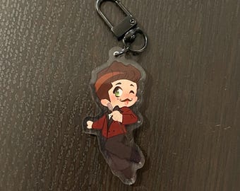 May include: A clear acrylic keychain featuring a cartoon illustration of a person with brown hair, a red jacket, and a black bow tie. The person is holding a microphone and has a mustache.