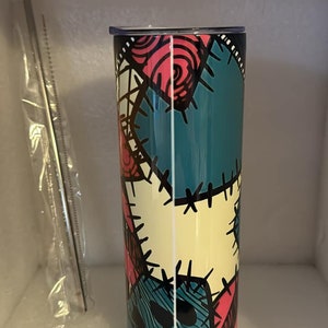 May include: A stainless steel tumbler with a colorful patchwork design featuring a stitched pattern and a character's face. The tumbler is teal, pink, yellow, and black.