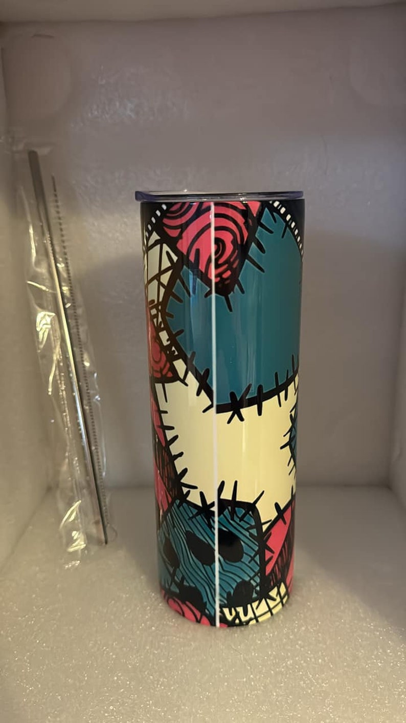 May include: A stainless steel tumbler with a colorful patchwork design featuring a stitched pattern and a character's face. The tumbler is teal, pink, yellow, and black.