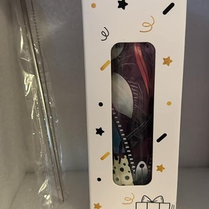 May include: A white box with a colorful design featuring a black and white gift box, stars, and confetti. The box contains a reusable straw with a colorful design.