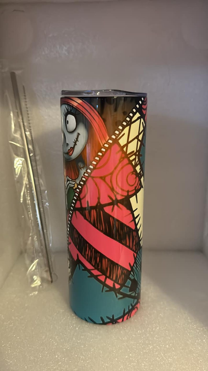 May include: A stainless steel tumbler with a colorful design featuring Sally from the Nightmare Before Christmas. The design includes pink, blue, black, and white colors.