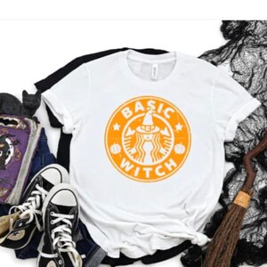 May include: White t-shirt with an orange graphic of a witch wearing a hat and a pumpkin. The graphic says "BASIC WITCH" in a circle.
