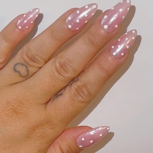 Può includere: Una mano con unghie lunghe a mandorla. Le unghie sono dipinte di rosa chiaro con pois bianchi. Le unghie hanno una finitura lucida. La mano ha un piccolo tatuaggio sul lato dell'indice.
