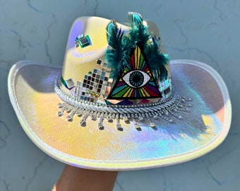 May include: A white cowboy hat with iridescent trim and a colorful eye patch with a triangle design. The hat is decorated with rhinestones and feathers.