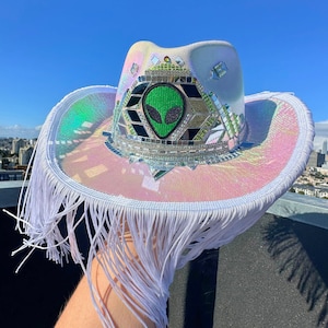 Könnte beinhalten: Ein weißer Cowboyhut mit Fransenbesatz und holografischem, schimmerndem Design. Der Hut hat einen grünen Alien-Gesichts-Patch in der Mitte.