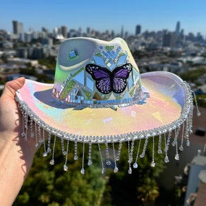 May include: Iridescent cowboy hat with a purple butterfly patch, mirror accents, and dangling crystal fringe. The hat has a pearlescent finish and a wide brim. A unique and eye-catching accessory.