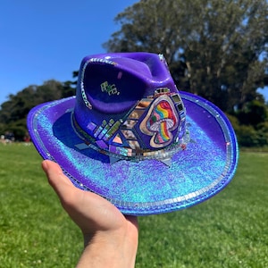 Puede incluir: Un sombrero de vaquero morado con un acabado holográfico iridiscente. El sombrero está decorado con detalles de espejo y un parche de hongo colorido. El sombrero se sostiene sobre un fondo de hierba verde y cielo azul.