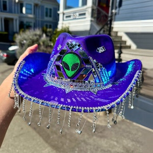 Puede incluir: Un sombrero vaquero morado con flecos plateados y un diseño de cara de extraterrestre verde. El sombrero está decorado con diamantes de imitación y azulejos espejados.