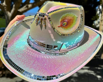 May include: A white cowboy hat with iridescent glitter and a small peacock feather design. The hat is decorated with small mirrored tiles.